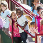 A big group of eight happy multiracial children sitting and standing together on the steps of playground equipment.  There are six girls and two boys of mixed ages from 2 to 8 years old, and of diverse ethnicities, shouting, laughing and smiling.