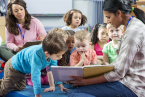 Female teacher giving a lesson to nursery students. They are sitting on the floor and there is a teacher taking notes.